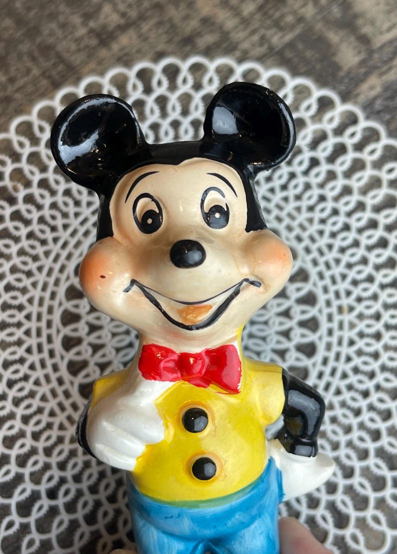 May include: A vintage ceramic figurine of Mickey Mouse. He is wearing a yellow shirt with two black buttons, a red bow tie, and blue pants. His face is painted with a friendly smile and large black eyes.
