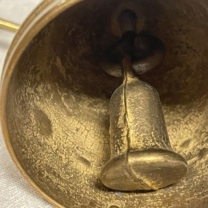 May include: Close-up of the inside of a brass bell with a worn, textured surface. The bell has a clapper inside that is also made of brass.