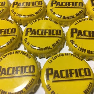 May include: A collection of yellow metal bottle caps featuring the brown text Pacifico, La Cerveza del Pacifico, and Año de 1900 Mazatlán, México. Each circular cap has a serrated edge and a glossy finish, displaying the brand name prominently in the center.