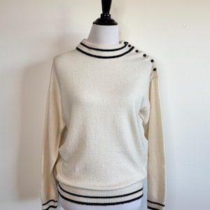 Puede incluir: Suéter color crema con cuello alto y mangas largas. El cuello, los puños y el dobladillo tienen rayas negras. Botones negros adornan el hombro. El suéter se exhibe en un maniquí.