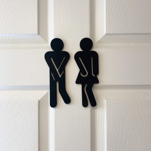 K&ouml;nnte beinhalten: Schwarz-wei&szlig;e Silhouetten-Schilder f&uuml;r eine Toilette, die eine Person mit gekreuzten Armen und eine Person mit gespreizten Beinen darstellen.