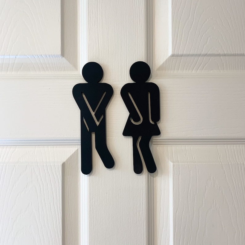 K&ouml;nnte beinhalten: Schwarz-wei&szlig;e Silhouetten-Schilder f&uuml;r eine Toilette, die eine Person mit gekreuzten Armen und eine Person mit gespreizten Beinen darstellen.
