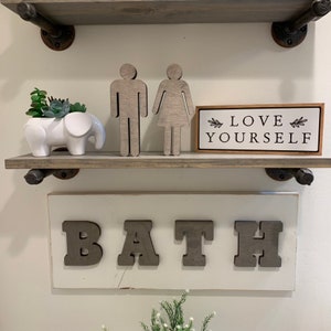 K&ouml;nnte beinhalten: Ein wei&szlig;es Holzschild mit dem Wort "BATH" in gro&szlig;en, grauen Holzbuchstaben. Das Schild ist an einer wei&szlig;en Wand &uuml;ber einem Regal mit einem kleinen wei&szlig;en Elefantentopf, einer Sukkulentenpflanze und zwei Holzfiguren, die einen Mann und eine Frau darstellen, angebracht. Ein kleines Holzschild mit dem Text "LOVE YOURSELF" befindet sich ebenfalls auf dem Regal.
