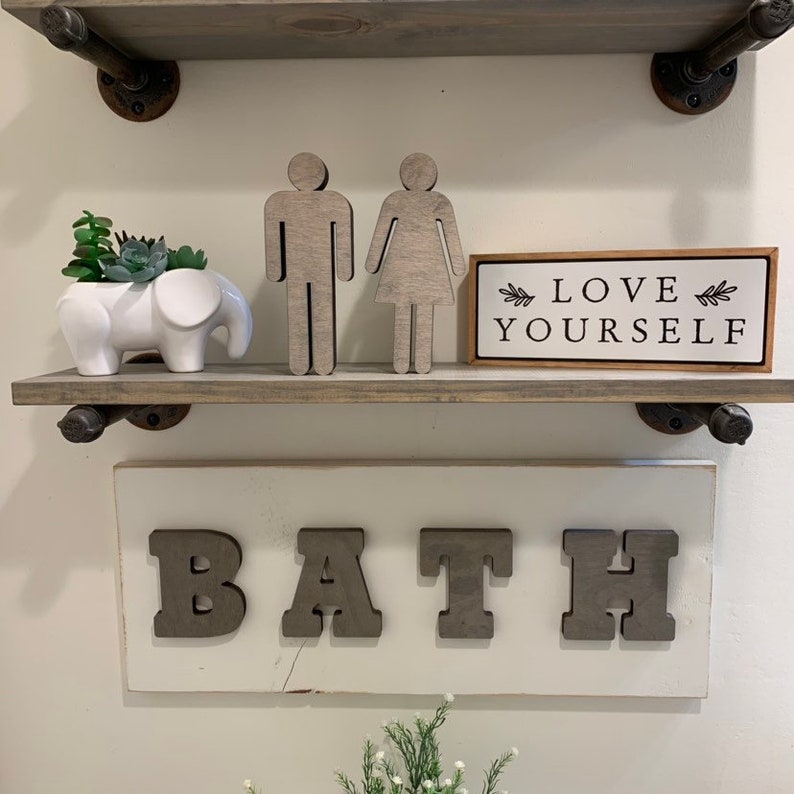 K&ouml;nnte beinhalten: Ein wei&szlig;es Holzschild mit dem Wort "BATH" in gro&szlig;en, grauen Holzbuchstaben. Das Schild ist an einer wei&szlig;en Wand &uuml;ber einem Regal mit einem kleinen wei&szlig;en Elefantentopf, einer Sukkulentenpflanze und zwei Holzfiguren, die einen Mann und eine Frau darstellen, angebracht. Ein kleines Holzschild mit dem Text "LOVE YOURSELF" befindet sich ebenfalls auf dem Regal.