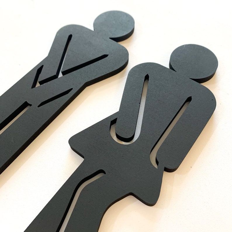 K&ouml;nnte beinhalten: Zwei schwarze Silhouette-Figuren, eine stehend mit verschr&auml;nkten Armen und die andere mit verschr&auml;nkten Armen und einem Rock, sind aus Holz ausgeschnitten.