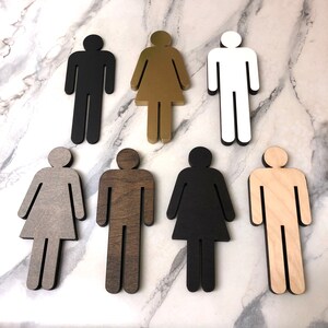 K&ouml;nnte beinhalten: Satz von sieben Toiletten Schildern aus Holz in verschiedenen Farben und Ausf&uuml;hrungen. Die Schilder sind in Form von Strichm&auml;nnchen gestaltet und zeigen eine Person und eine Frau.