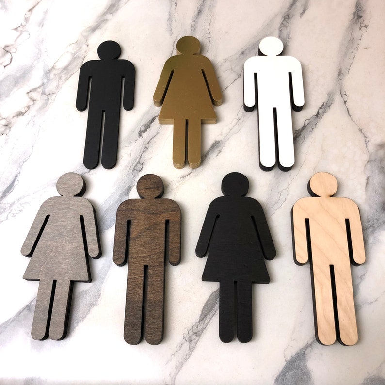 K&ouml;nnte beinhalten: Satz von sieben Toiletten Schildern aus Holz in verschiedenen Farben und Ausf&uuml;hrungen. Die Schilder sind in Form von Strichm&auml;nnchen gestaltet und zeigen eine Person und eine Frau.