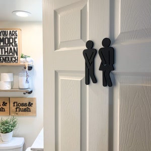 K&ouml;nnte beinhalten: Schwarze Silhouetten von Toilettenzeichen f&uuml;r M&auml;nner und Frauen an einer wei&szlig;en T&uuml;r. Die Schilder sind in Form von Strichm&auml;nnchen mit den H&auml;nden auf den H&uuml;ften.