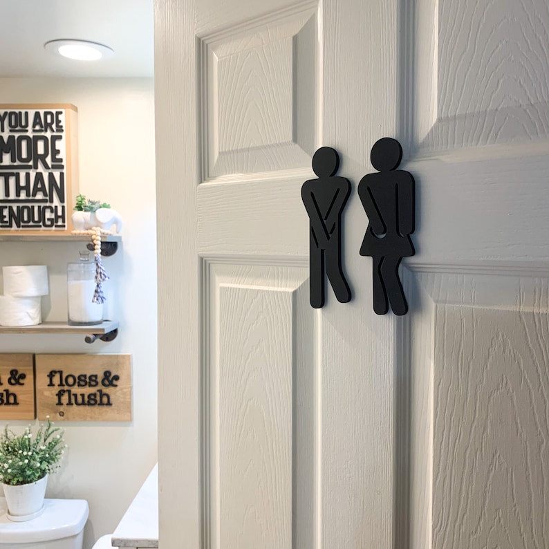 K&ouml;nnte beinhalten: Schwarze Silhouetten von Toilettenzeichen f&uuml;r M&auml;nner und Frauen an einer wei&szlig;en T&uuml;r. Die Schilder sind in Form von Strichm&auml;nnchen mit den H&auml;nden auf den H&uuml;ften.