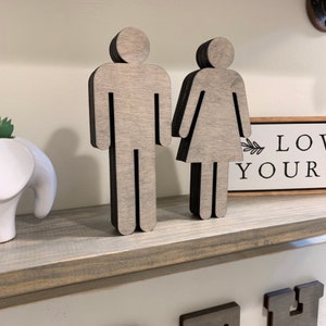 K&ouml;nnte beinhalten: Zwei ausgeschnittene Holzfiguren einer Person, eine mit einem Rock und die andere mit einer Hose, stehen auf einem grauen Regal. Die Figuren sind in hellgrau mit schwarzen Umrissen bemalt. Ein kleines Holzschild mit dem Text "Love Your" ist im Hintergrund sichtbar.