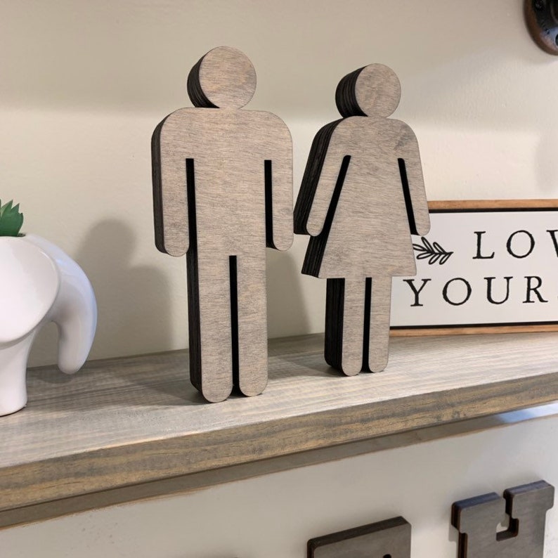 K&ouml;nnte beinhalten: Zwei ausgeschnittene Holzfiguren einer Person, eine mit einem Rock und die andere mit einer Hose, stehen auf einem grauen Regal. Die Figuren sind in hellgrau mit schwarzen Umrissen bemalt. Ein kleines Holzschild mit dem Text "Love Your" ist im Hintergrund sichtbar.