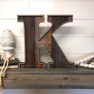 May include: A large wooden letter K with a dark brown wood grain and a light brown wood grain. The letter is framed with a dark brown wood frame. The letter is sitting on a gray wooden shelf.