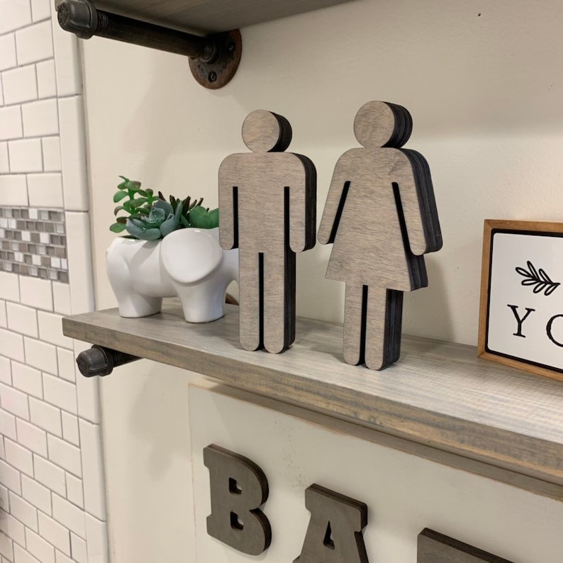 K&ouml;nnte beinhalten: Ein Holzregal mit zwei grauen Holzfiguren, eine m&auml;nnliche und eine weibliche, die ein Toilettenzeichen darstellen. Das Regal ist aus wiederverwendetem Holz gefertigt und hat einen rustikalen Look. Die Figuren sind aus Holz ausgeschnitten und haben ein schlichtes Design. Das Regal ist an einer wei&szlig;en Wand montiert.