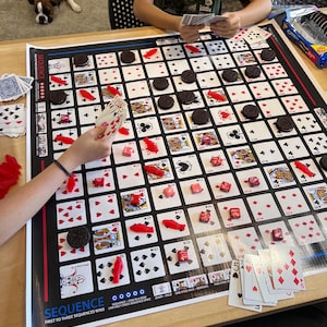 May include: A game of Sequence is in progress. The game board is black with red and white playing cards. Red candy pieces are placed on the board. The game instructions are visible at the bottom of the board. The text reads "SEQUENCE - FIVE IN A ROW - FIRST TO THREE SEQUENCES WINS - CAN ONLY CROSS SEQUENCE ONCE - JOKER - WILD - ONE EYED JACK REMOVES - TWO EYED JACK - REMOVE TWO".