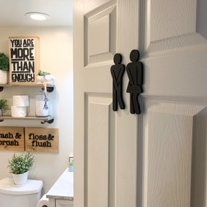 K&ouml;nnte beinhalten: Schwarze Holzsilhouette eines Mannes und einer Frau, die nebeneinander auf einer wei&szlig;en T&uuml;r stehen. Die Silhouetten sind stilisiert, um wie Personen auszusehen, die die Toilette benutzen. Die T&uuml;r hat eine wei&szlig;e Lackierung mit Holzmaserung.