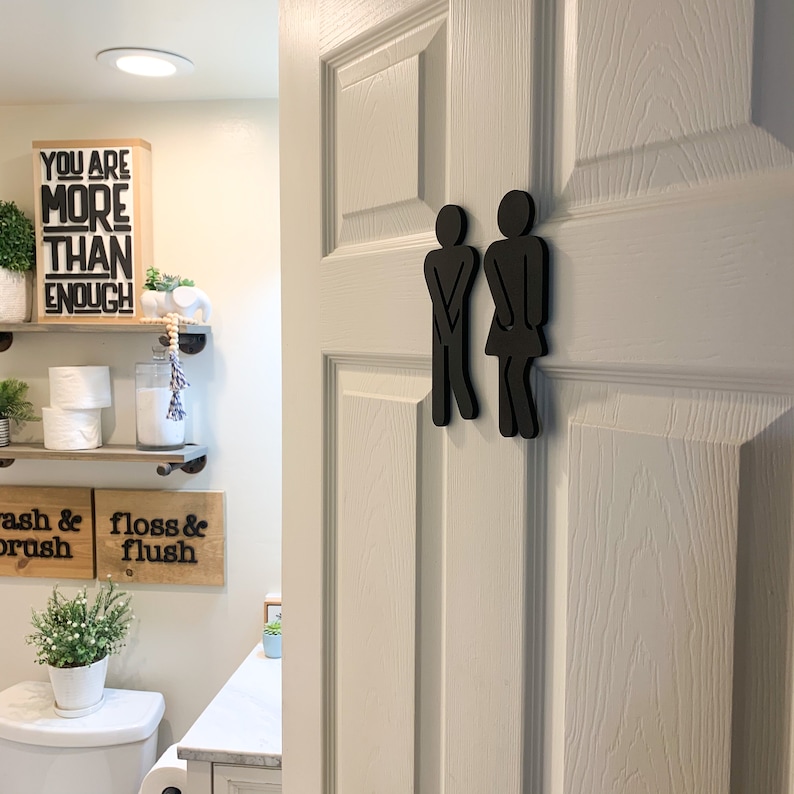 K&ouml;nnte beinhalten: Schwarze Holzsilhouette eines Mannes und einer Frau, die nebeneinander auf einer wei&szlig;en T&uuml;r stehen. Die Silhouetten sind stilisiert, um wie Personen auszusehen, die die Toilette benutzen. Die T&uuml;r hat eine wei&szlig;e Lackierung mit Holzmaserung.