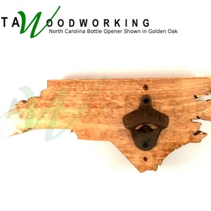 May include: A wooden bottle opener shaped like the state of North Carolina. The opener is made of natural wood with a rustic finish. The bottle opener is mounted on a wall. The text "KATA WOODWORKING" is visible in the top left corner of the image. The text "North Carolina Bottle Opener Shown in Golden Oak" is visible below the text "KATA WOODWORKING".