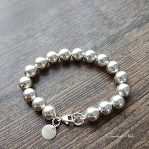 Può includere: Un braccialetto d'argento con grandi perline rotonde. Il braccialetto è realizzato in metallo lucido e ha una chiusura sicura con un piccolo ciondolo rotondo. Le perline sono uniformemente distanziate e riflettono la luce, creando un aspetto lucido. Il braccialetto è esposto su una superficie di legno.