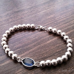 Puede incluir: Pulsera de plata con cuentas esféricas y una gema ovalada azul oscuro. La pulsera tiene un cierre seguro. La gema está engastada en un bisel de plata. La pulsera se muestra sobre una superficie de madera oscura.