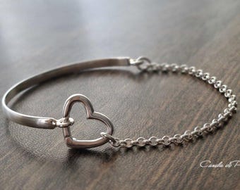 May include: A silver bracelet with a heart-shaped charm. The bracelet features a solid bar on one side and a delicate chain on the other, connected by the heart. The bracelet is displayed on a dark wooden surface.