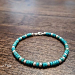 Op de afbeelding: Een turquoise kralenarmband met zilverkleurige accenten. De armband heeft kleine, ronde turquoise kralen afgewisseld met grotere, zilverkleurige kralen. De sluiting is ook zilverkleurig. De armband wordt getoond op een donker houten oppervlak.