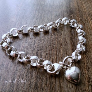 Peut inclure: Un bracelet en argent avec un charm en forme de cœur. Le bracelet est composé d'une chaîne de maillons arrondis interconnectés. Un petit charm en forme de cœur poli est suspendu à la fermeture. Le bracelet est présenté sur une surface en bois.
