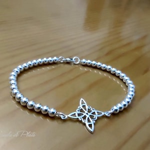 May include: A silver bracelet featuring a beaded chain and a decorative four-pointed star charm. The charm has an openwork design. The bracelet is displayed on a light brown wooden surface.