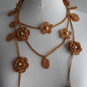 May include: A long, multi-strand crocheted necklace with brown flowers and pearl accents. The necklace is made of a mustard yellow yarn and features a delicate floral design.