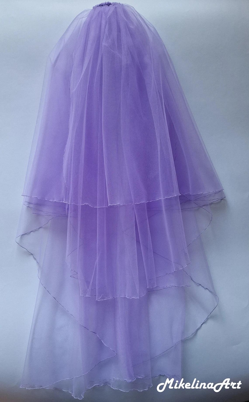 May include: A layered, lavender bridal veil. The veil is made of sheer fabric and features multiple tiers, creating a flowing effect. The bottom edges are gently scalloped. The Mikellina Art logo is visible in the bottom right corner.