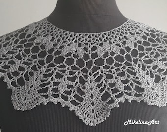 May include: A silver crochet collar with a delicate floral pattern. The collar is made of fine thread and has a scalloped edge.