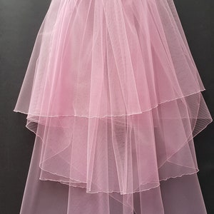 May include: A layered, light pink bridal veil. The veil is made of sheer, delicate fabric, with multiple tiers cascading down. The veil is gathered at the top, creating a full, flowing effect. The bottom of the veil is slightly scalloped.