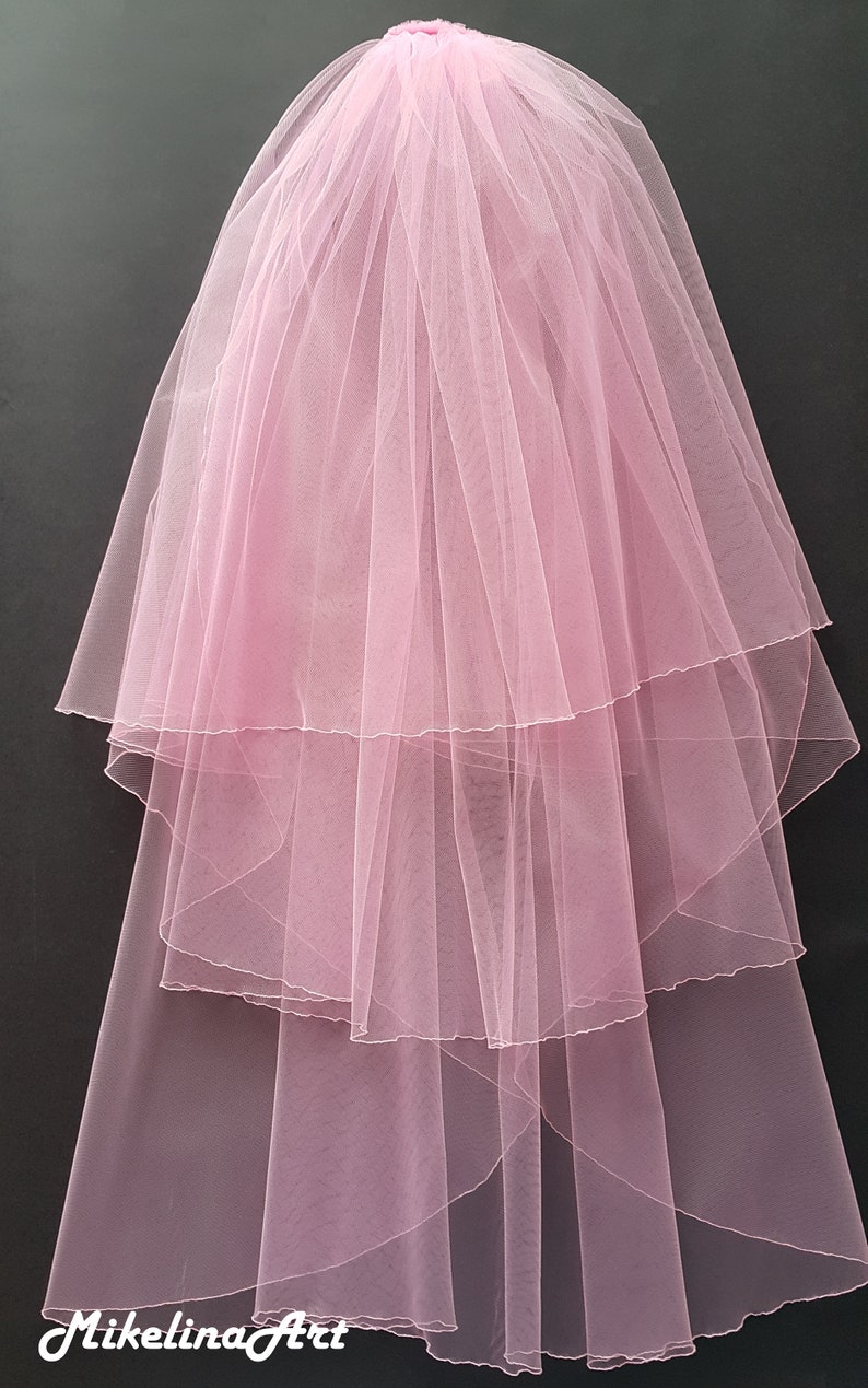 May include: A layered, light pink bridal veil. The veil is made of sheer, delicate fabric, with multiple tiers cascading down. The veil is gathered at the top, creating a full, flowing effect. The bottom of the veil is slightly scalloped.