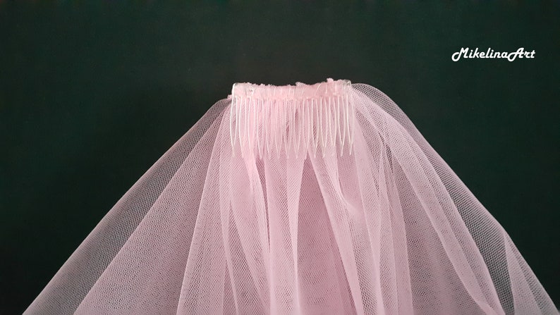 May include: A light pink bridal veil with a comb. The veil is made of a sheer, delicate fabric and is attached to a plastic comb. The text "MikelinaArt" is in the upper right corner. The background is a dark green.