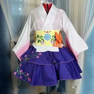 Puede incluir: Un disfraz de estilo japonés exhibido en un maniquí. El atuendo presenta una parte superior blanca con detalles en rosa, una faja amarilla con bordados florales y una falda morada en capas con detalles florales. Un lazo naranja añade un toque de color.