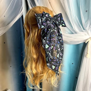 May include: A black hair bow with a celestial pattern of moons, stars, and potion bottles. The bow is tied on a blonde wig. The background includes sheer white and blue curtains.