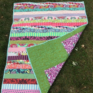 May include: A colorful patchwork quilt with a green backing. The quilt is made of strips of fabric in various patterns and colors, including pink, red, blue, green, and purple. The quilt is folded in half and lying on a bed of green grass.