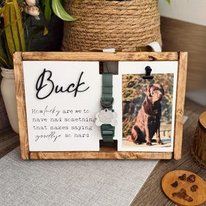May include: A wooden framed pet memorial with a photo of a brown dog, a green collar, and a name tag. The name "Buck" is displayed in cursive, with a quote below. A paw print coaster is also visible.