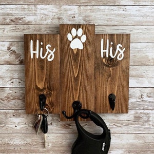 May include: A wooden key holder with two hooks and the words "His" on either side of a paw print. A set of keys and a black retractable dog leash are hanging on the hooks.