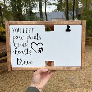 May include: A wooden sign with a white background and black text that reads "You left paw prints on our hearts Bruce". The sign has a heart with a paw print inside and a small clip at the top.