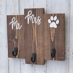 Op de afbeelding: Een houten sleutelhanger met drie haken, geschilderd in bruin. De woorden "Mr" en "Mrs" zijn in wit geschilderd op het middelste gedeelte. Een witte pootafdruk is geschilderd op het rechter gedeelte.