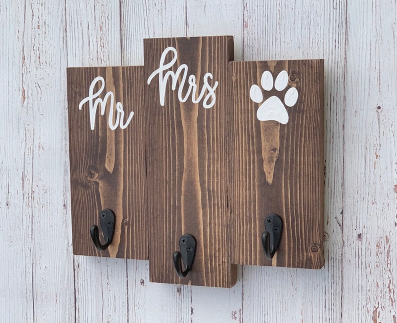 Op de afbeelding: Een houten sleutelhanger met drie haken, geschilderd in bruin. De woorden "Mr" en "Mrs" zijn in wit geschilderd op het middelste gedeelte. Een witte pootafdruk is geschilderd op het rechter gedeelte.