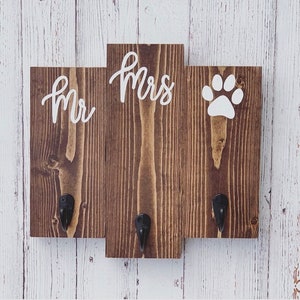 Op de afbeelding: Een houten bord met drie secties, elk met een wit ontwerp. De linker sectie zegt "Mr." in cursief, de middelste sectie zegt "Mrs." in cursief, en de rechter sectie heeft een witte voetafdruk. Elke sectie heeft een zwarte metalen haak.