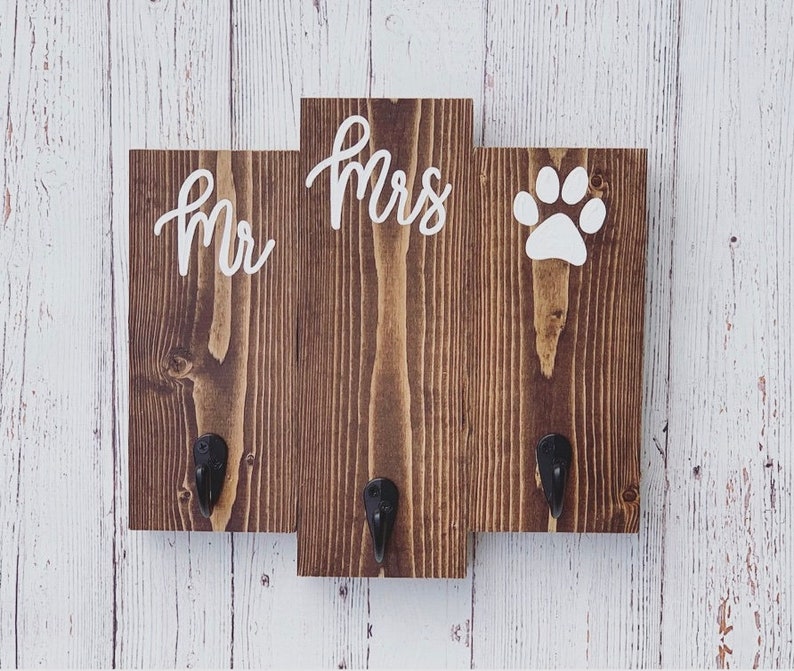 Op de afbeelding: Een houten bord met drie secties, elk met een wit ontwerp. De linker sectie zegt "Mr." in cursief, de middelste sectie zegt "Mrs." in cursief, en de rechter sectie heeft een witte voetafdruk. Elke sectie heeft een zwarte metalen haak.