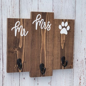 Op de afbeelding: Een houten bord met de woorden "Mr." en "Mrs." en een pootprint in witte verf. Het bord heeft drie haken om spullen aan op te hangen.