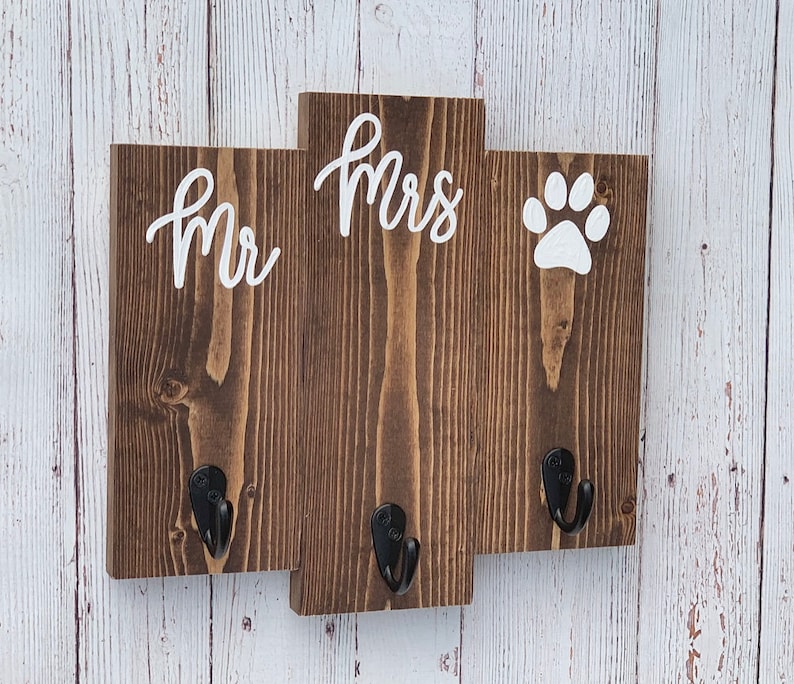 Op de afbeelding: Een houten bord met de woorden "Mr." en "Mrs." en een pootprint in witte verf. Het bord heeft drie haken om spullen aan op te hangen.