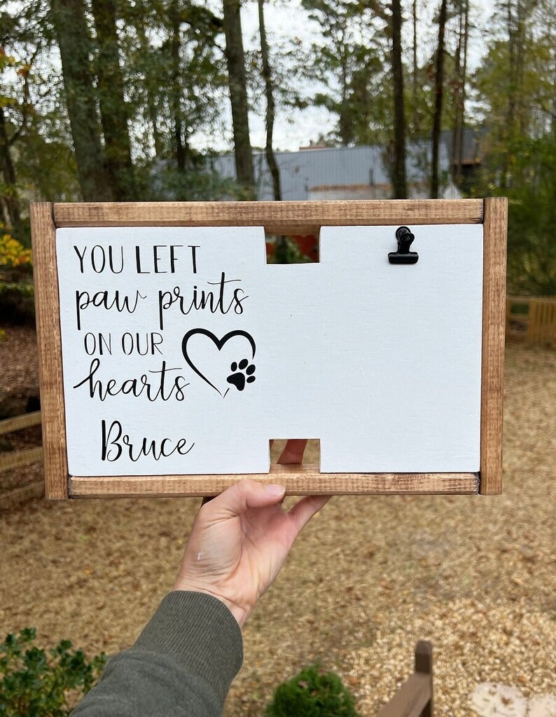May include: A wooden sign with a white background and black text that reads "You left paw prints on our hearts Bruce". The sign has a heart with a paw print inside and a small clip at the top.