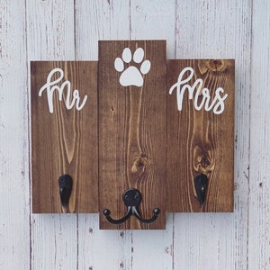 Op de afbeelding: Een houten bord met de woorden "Mr." en "Mrs." aan weerszijden van een pootprint. Het bord heeft twee zwarte haken aan de onderkant.