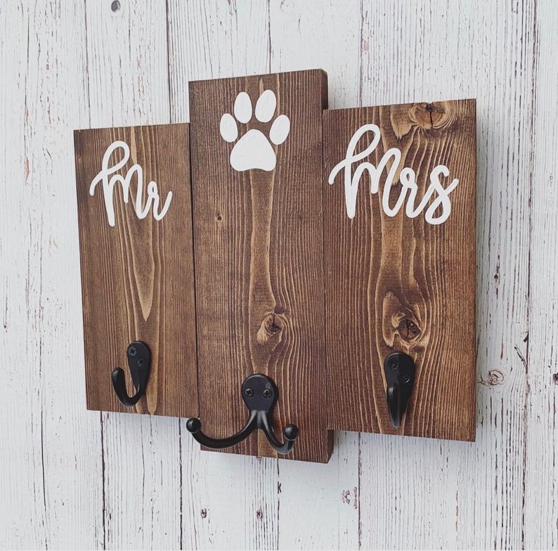 Op de afbeelding: Een houten bord met de woorden "Mr." en "Mrs." aan weerszijden van een pootprint. Het bord heeft twee zwarte metalen haken.