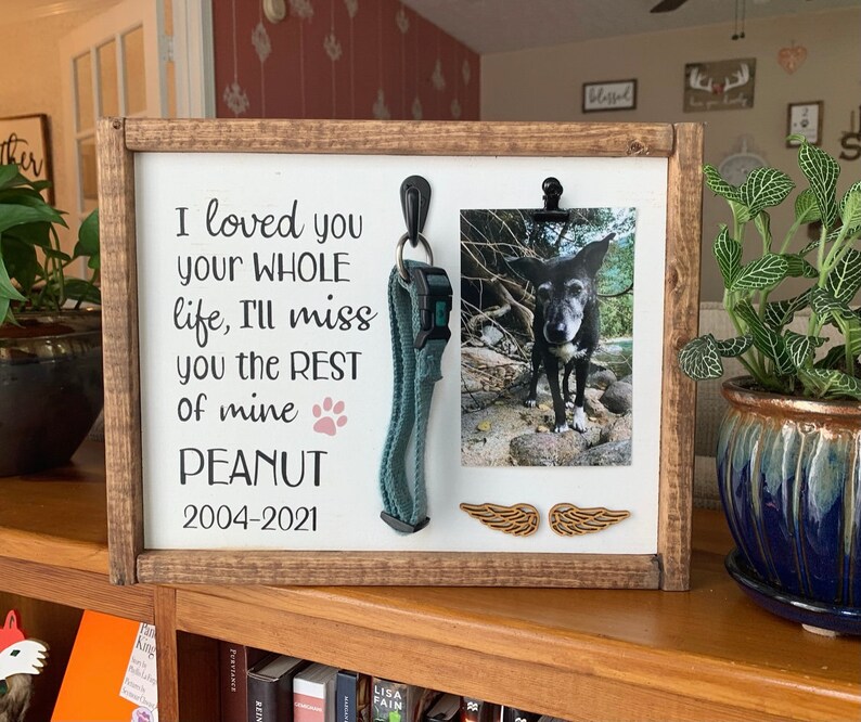 May include: A wooden memorial plaque with a photo of a black and white dog, a dog collar, and two wooden angel wings. The plaque reads "I loved you your whole life, I'll miss you the rest of mine PEANUT 2004-2021".