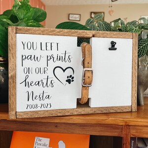 May include: A wooden sign with the text "You left paw prints on our hearts Nesta 2008-2023". A heart with paw prints is in the center of the sign. A brown leather dog collar is attached to the sign with a black clip.